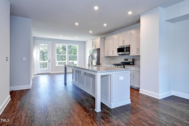 kitchen with dark wood-style flooring, stainless steel appliances, baseboards, decorative backsplash, and a kitchen island with sink