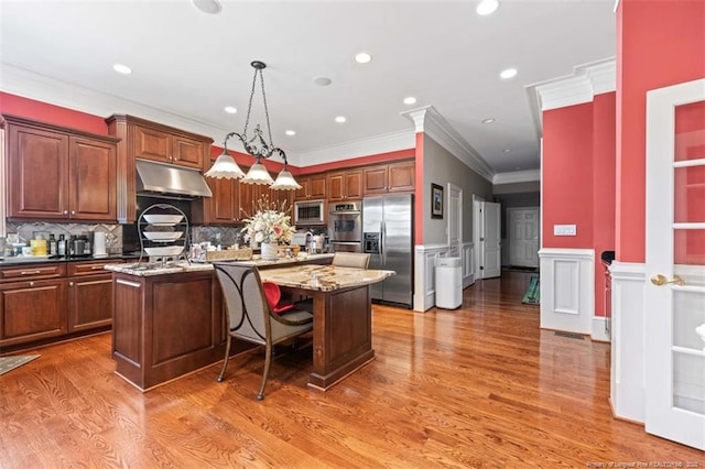 kitchen featuring a wainscoted wall, under cabinet range hood, stainless steel appliances, a kitchen island with sink, and light wood-style floors