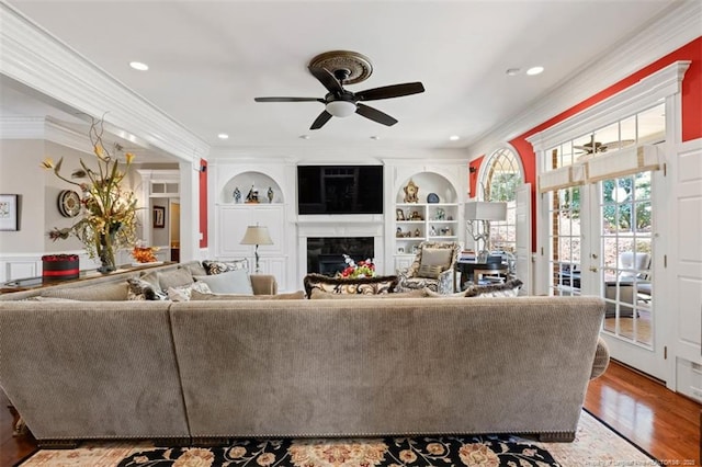 living room featuring built in shelves, a fireplace, wood finished floors, ceiling fan, and ornamental molding
