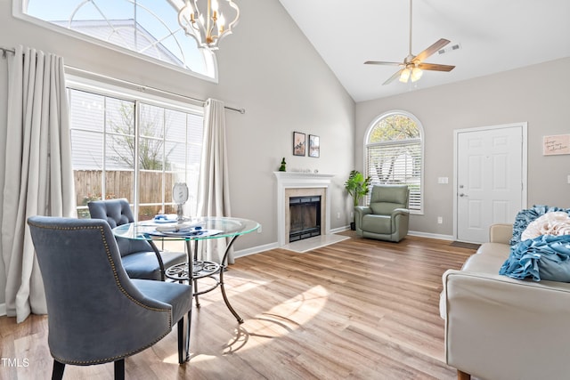 living area with ceiling fan with notable chandelier, high vaulted ceiling, baseboards, light wood finished floors, and a tiled fireplace
