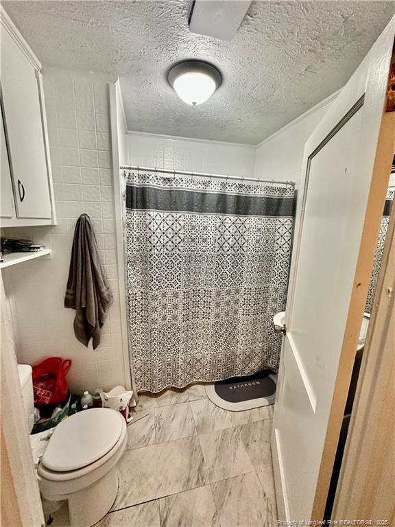 full bath featuring toilet, a shower with curtain, a textured ceiling, marble finish floor, and tile walls