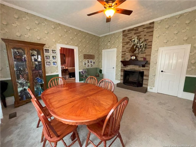 carpeted dining space featuring ceiling fan, wallpapered walls, crown molding, and a stone fireplace