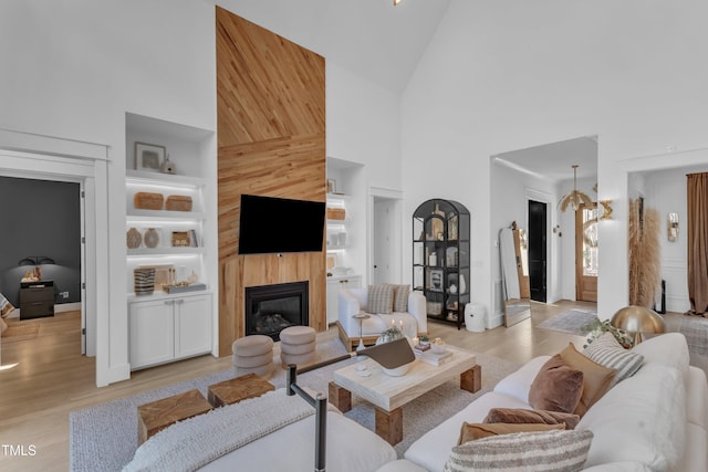 living area featuring high vaulted ceiling, light wood-style flooring, a glass covered fireplace, and built in shelves