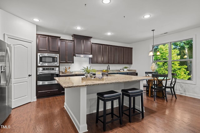 kitchen featuring stainless steel appliances, dark brown cabinetry, a kitchen island with sink, light stone countertops, and dark wood-style flooring