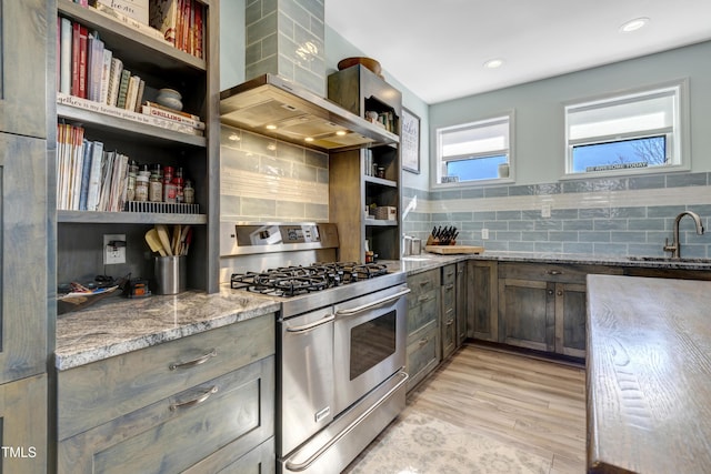 kitchen with wall chimney range hood, open shelves, light stone counters, a sink, and range with two ovens