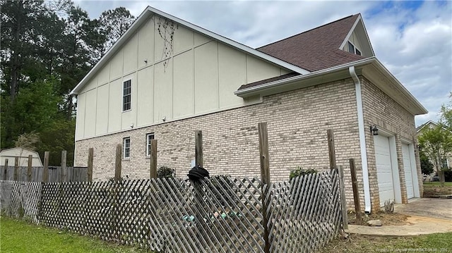 view of side of property with roof with shingles, driveway, an attached garage, brick siding, and fence