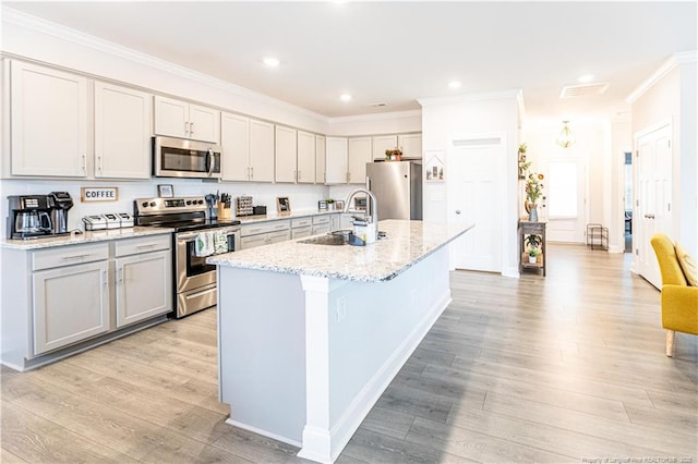 kitchen with light wood-style flooring, ornamental molding, appliances with stainless steel finishes, a sink, and a center island with sink