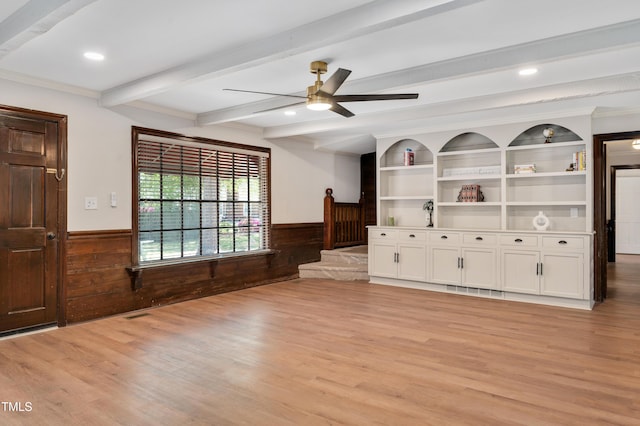 unfurnished living room featuring light wood-style flooring, a ceiling fan, wood walls, wainscoting, and beam ceiling