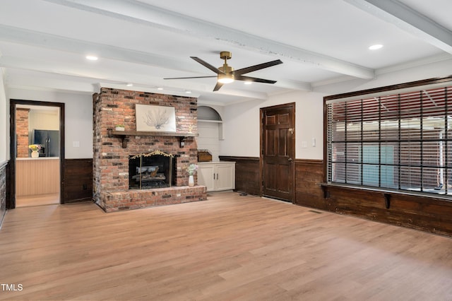 unfurnished living room featuring a fireplace, light wood-style flooring, a ceiling fan, wainscoting, and beam ceiling