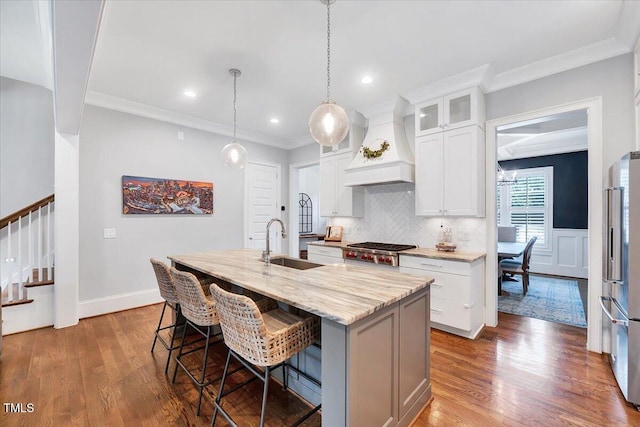 kitchen with custom exhaust hood, a sink, high quality fridge, a kitchen island with sink, and range