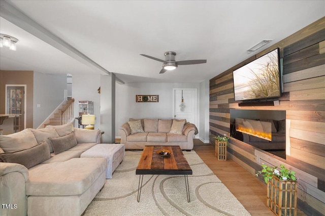 living area with a glass covered fireplace, wood finished floors, ceiling fan, and stairs