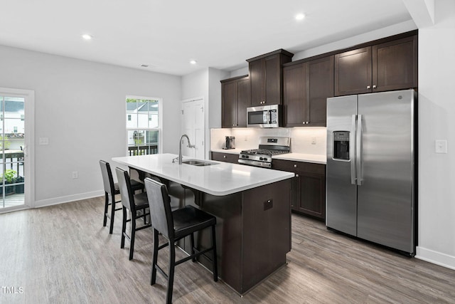 kitchen with stainless steel appliances, dark brown cabinets, wood finished floors, a sink, and a center island with sink