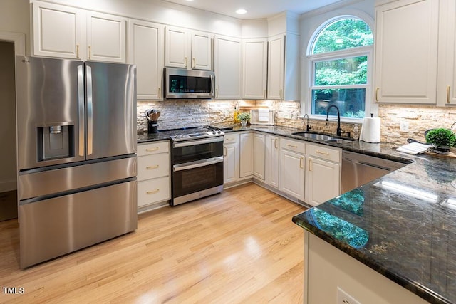 kitchen featuring appliances with stainless steel finishes, a sink, dark stone counters, light wood finished floors, and backsplash