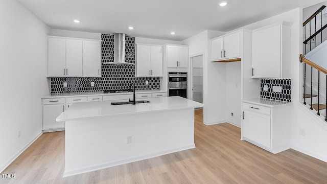 kitchen featuring wall chimney range hood, a sink, double oven, light wood-type flooring, and decorative backsplash