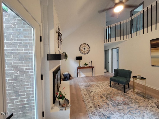 living area featuring wood finished floors, ceiling fan, high vaulted ceiling, a glass covered fireplace, and baseboards