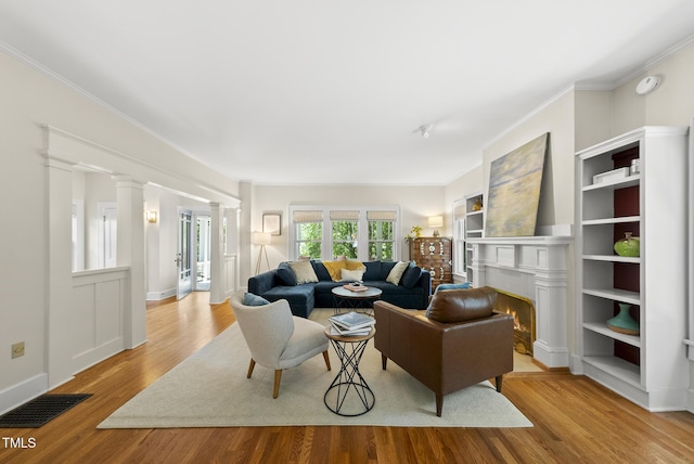 living area with visible vents, crown molding, wood finished floors, a warm lit fireplace, and ornate columns