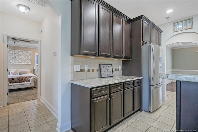 kitchen with arched walkways, freestanding refrigerator, dark brown cabinetry, backsplash, and light tile patterned flooring