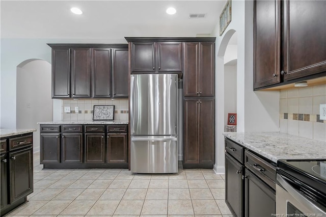 kitchen featuring light tile patterned floors, arched walkways, freestanding refrigerator, dark brown cabinets, and light stone countertops