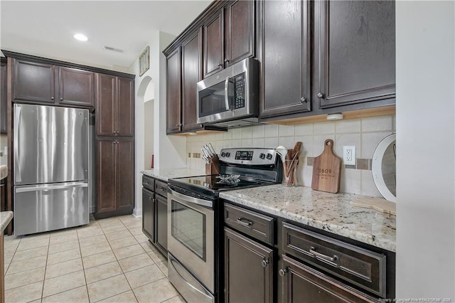kitchen featuring dark brown cabinetry, arched walkways, light tile patterned flooring, stainless steel appliances, and decorative backsplash