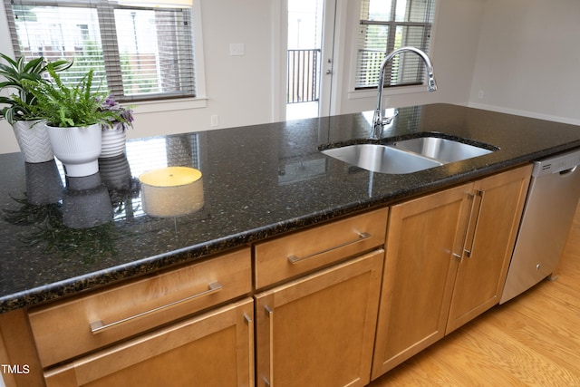 kitchen with light wood-style flooring, brown cabinets, dark stone counters, stainless steel dishwasher, and a sink