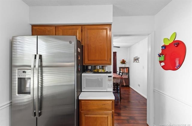 kitchen featuring white microwave, stainless steel fridge with ice dispenser, brown cabinetry, dark wood-style flooring, and light countertops