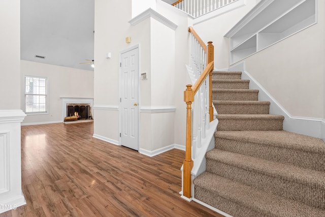 staircase featuring visible vents, baseboards, ceiling fan, wood finished floors, and a lit fireplace