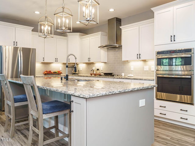 kitchen featuring wall chimney exhaust hood, stainless steel appliances, white cabinets, light wood-type flooring, and an island with sink
