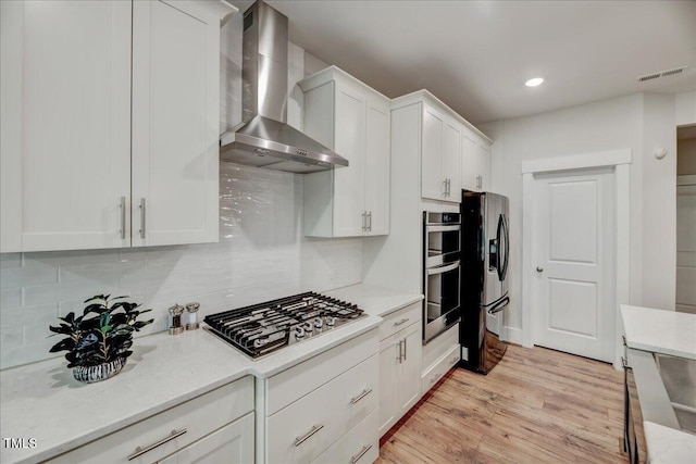 kitchen featuring stainless steel appliances, wall chimney range hood, light wood-style flooring, tasteful backsplash, and white cabinetry