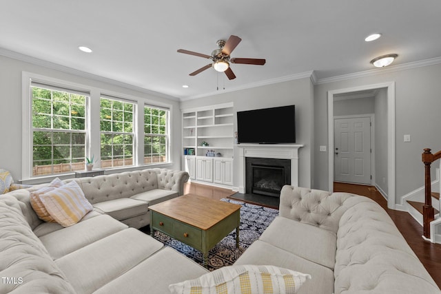 living area featuring ornamental molding, a ceiling fan, built in shelves, a fireplace with flush hearth, and dark wood-style flooring