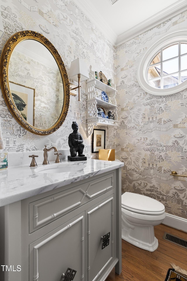 bathroom with wallpapered walls, wood finished floors, crown molding, vanity, and toilet