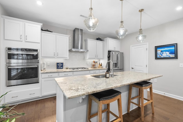 kitchen with wall chimney range hood, appliances with stainless steel finishes, a center island with sink, a sink, and a breakfast bar
