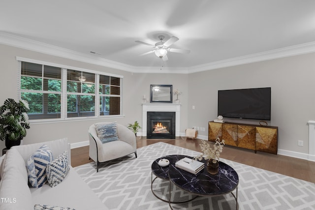 living room with crown molding, wood finished floors, ceiling fan, a fireplace with flush hearth, and baseboards