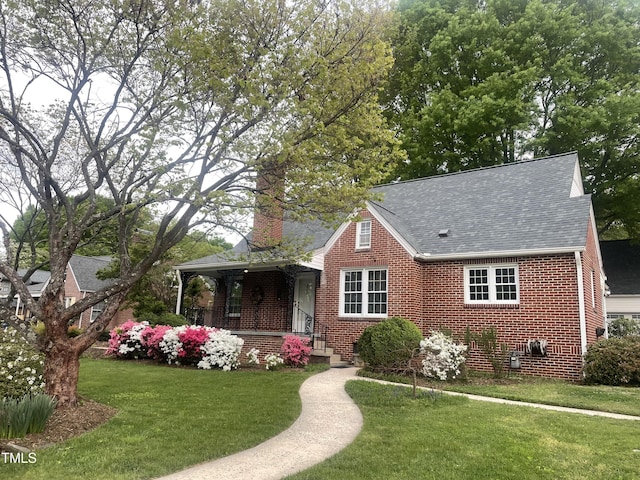view of front facade with a chimney, a front yard, brick siding, a porch, and a shingled roof