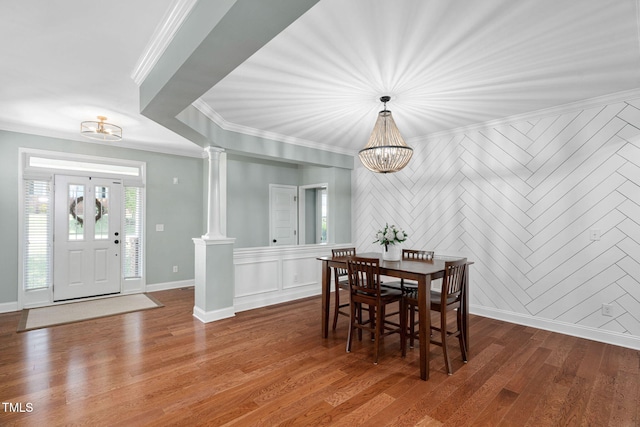 dining space featuring wood finished floors, a notable chandelier, ornamental molding, a decorative wall, and ornate columns