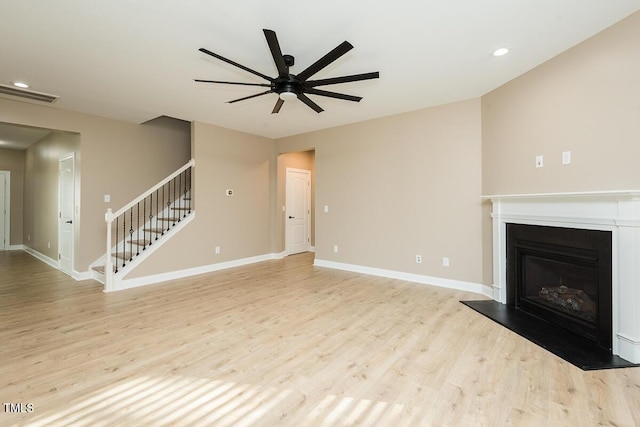 unfurnished living room with light wood-style flooring, a glass covered fireplace, stairs, and ceiling fan
