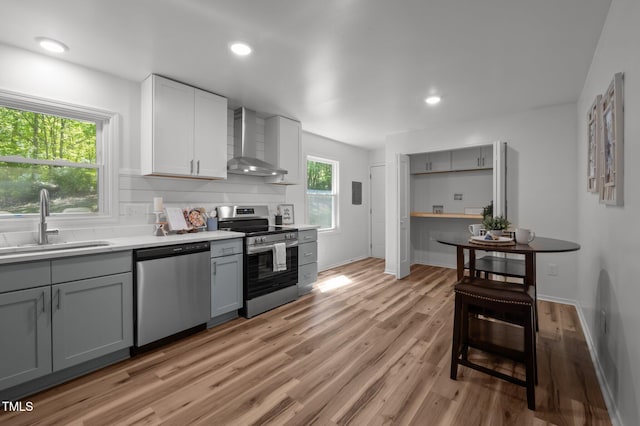 kitchen with stainless steel appliances, gray cabinets, a sink, light wood-type flooring, and wall chimney exhaust hood