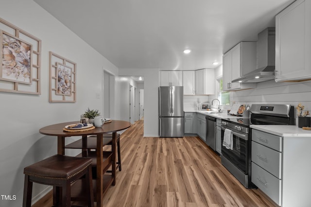 kitchen with a sink, light wood-style floors, wall chimney range hood, light countertops, and appliances with stainless steel finishes