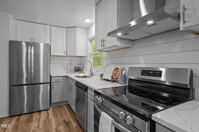 kitchen featuring wall chimney range hood, light stone counters, light wood-type flooring, white cabinets, and appliances with stainless steel finishes