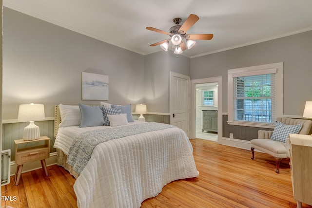 bedroom with light wood-type flooring, crown molding, a wainscoted wall, connected bathroom, and a ceiling fan