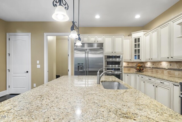 kitchen with decorative backsplash, glass insert cabinets, light stone counters, a sink, and stainless steel appliances