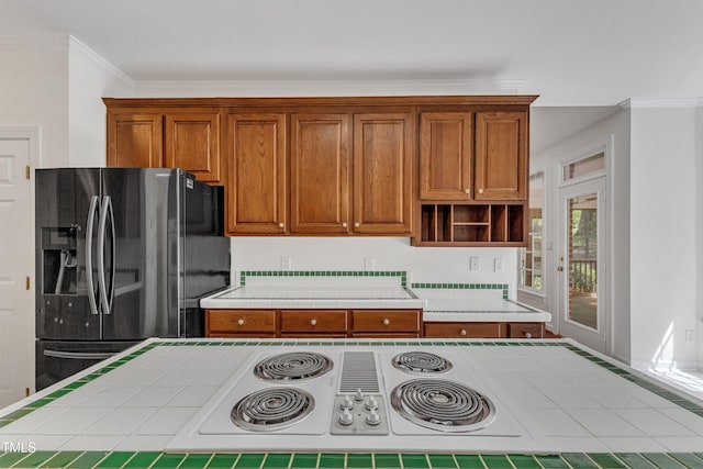 kitchen with crown molding, open shelves, brown cabinetry, tile counters, and refrigerator with ice dispenser