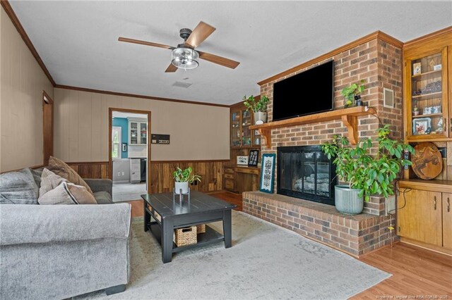 living area featuring wood finished floors, crown molding, ceiling fan, wooden walls, and a brick fireplace