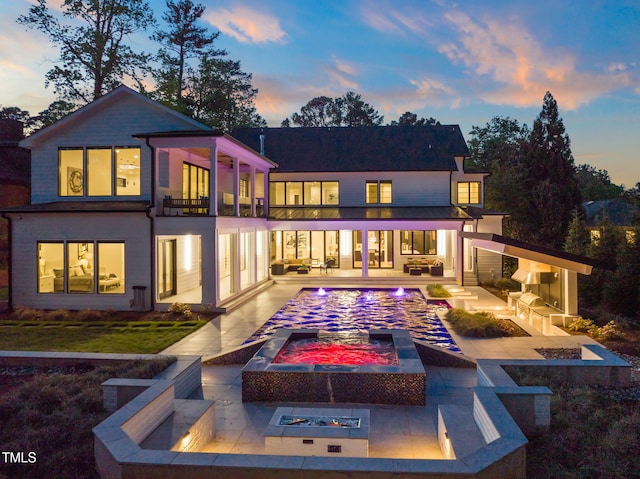 back of property at dusk featuring an outdoor living space with a fire pit, an outdoor pool, an in ground hot tub, a patio area, and a balcony