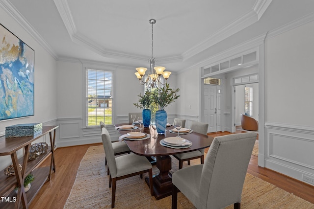dining space featuring an inviting chandelier, a raised ceiling, wood finished floors, ornamental molding, and a decorative wall