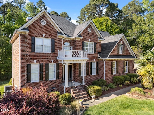 colonial inspired home featuring a balcony, a front lawn, cooling unit, brick siding, and roof with shingles