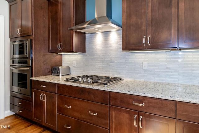 kitchen featuring stainless steel appliances, light stone countertops, wall chimney exhaust hood, and light wood-type flooring