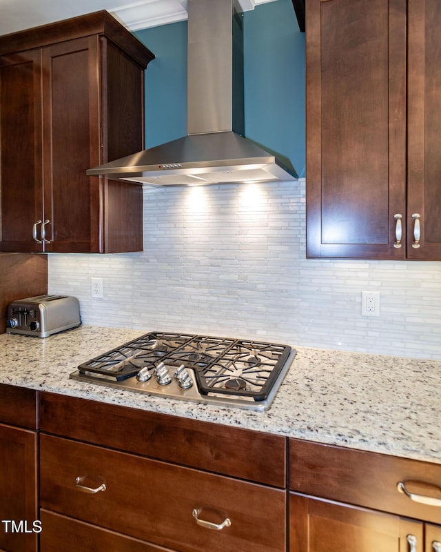 kitchen featuring dark brown cabinetry, wall chimney exhaust hood, light stone counters, backsplash, and stainless steel gas stovetop