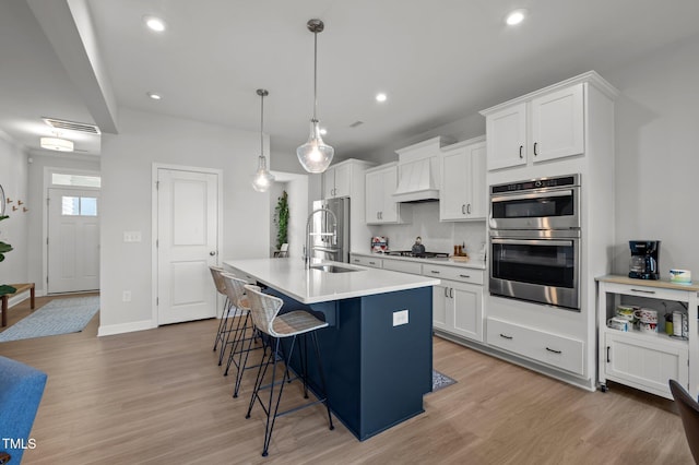 kitchen featuring custom exhaust hood, an island with sink, a sink, light wood-style flooring, and appliances with stainless steel finishes