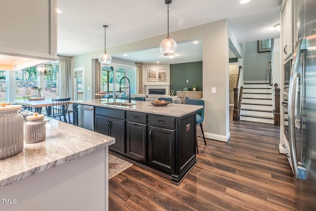 kitchen with a kitchen island with sink, a sink, dark wood-style flooring, stainless steel appliances, and decorative light fixtures