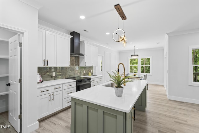 kitchen featuring an island with sink, wall chimney range hood, electric range oven, white cabinetry, and a sink